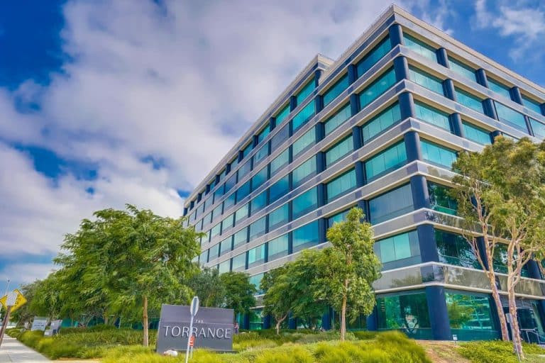 A modern, multi-story office building with large windows, surrounded by trees. A sign in front reads "Torrance." The sky is partly cloudy.